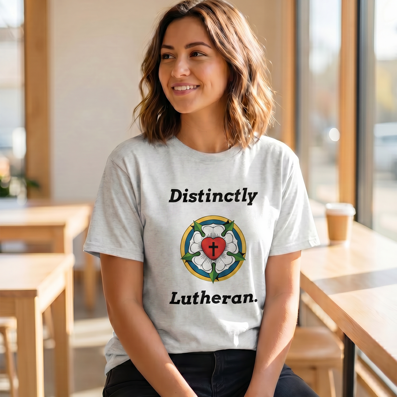A smiling woman wearing a white t-shirt featuring the Luther Rose seal and the text "Distinctly Lutheran" in a modern cafe setting. Perfect graphic tee for Lutheran apparel and Christian merchandise.