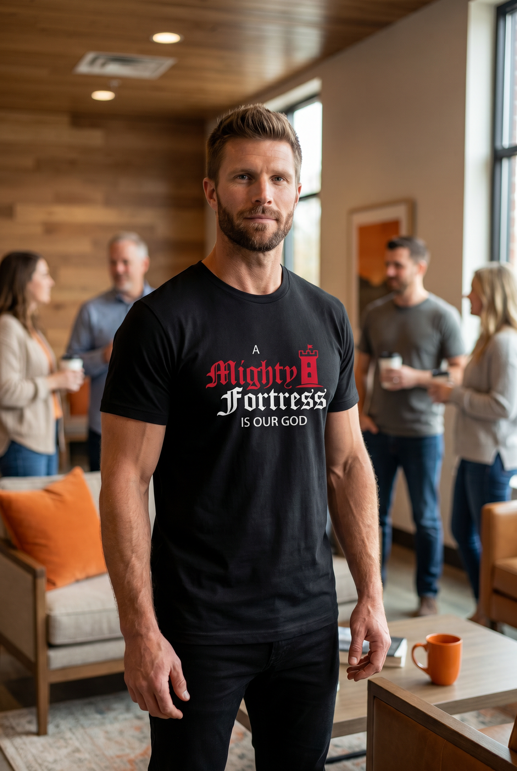 A man standing in a church fellowship hall wearing a black Christian graphic tee with the words 'A Mighty Fortress Is Our God' in bold Gothic script. Traditional Lutheran clothing from Phoenix Anthem.