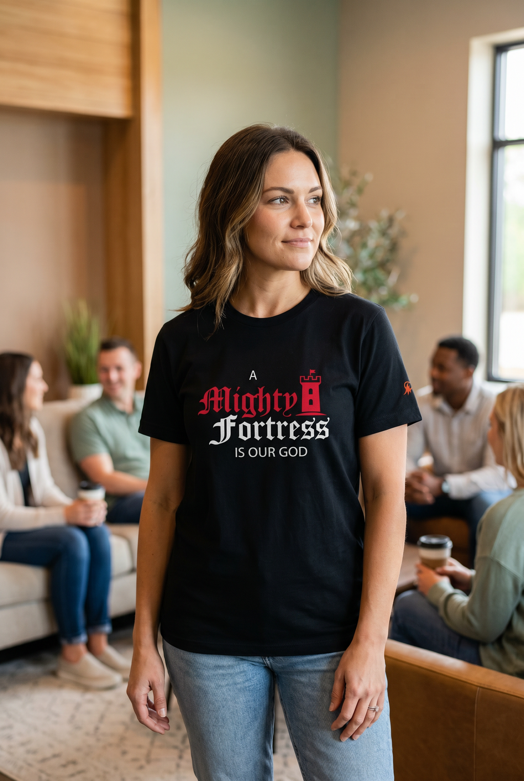 Smiling woman standing in a church common room wearing a black 'A Mighty Fortress Is Our God' shirt featuring a red castle tower graphic. Reformed faith-based apparel from Phoenix Anthem.
