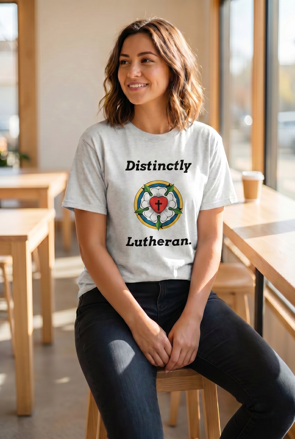 Smiling woman sitting on a stool in a sunlit coffee shop wearing a white 'Distinctly Lutheran' graphic tee featuring the traditional Luther Rose seal. Lifestyle Christian apparel from Phoenix Anthem.