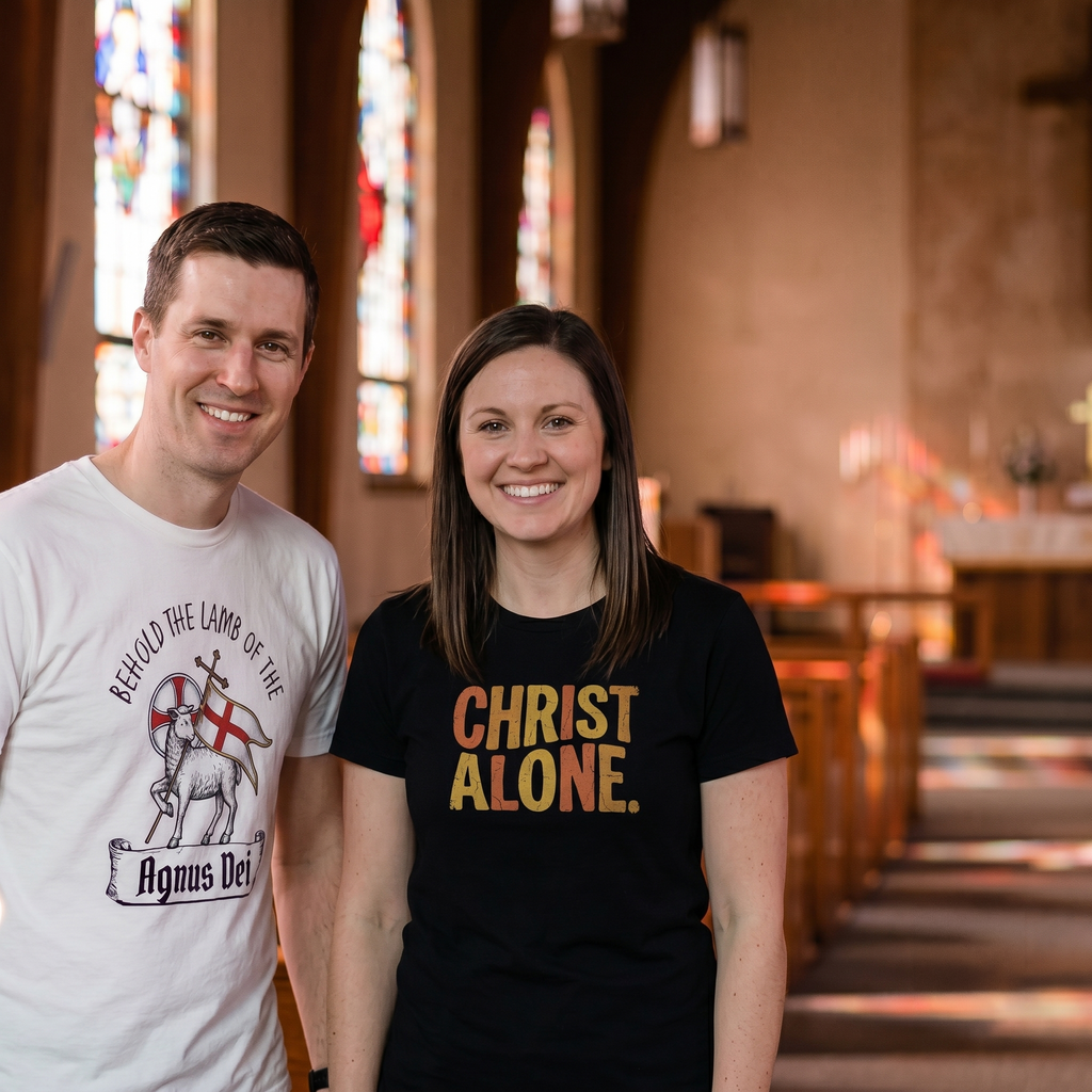 Smiling Christian couple standing together inside a traditional church sanctuary with pews and stained glass windows. The man is wearing a white 'Behold the Lamb of God' Agnus Dei tee, and the woman is wearing a black retro 'Christ Alone' shirt. Coordinated Lutheran lifestyle apparel from Phoenix Anthem.