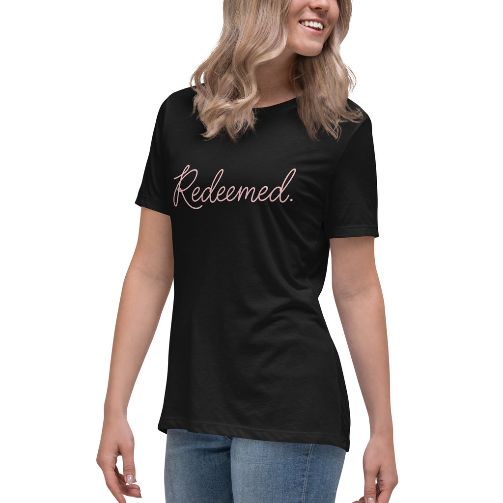 Three-quarter front left view of a blonde woman wearing a black relaxed-fit Christian t-shirt displaying the full typography design 'Redeemed.' in an elegant pink script. Lutheran women's fashion.