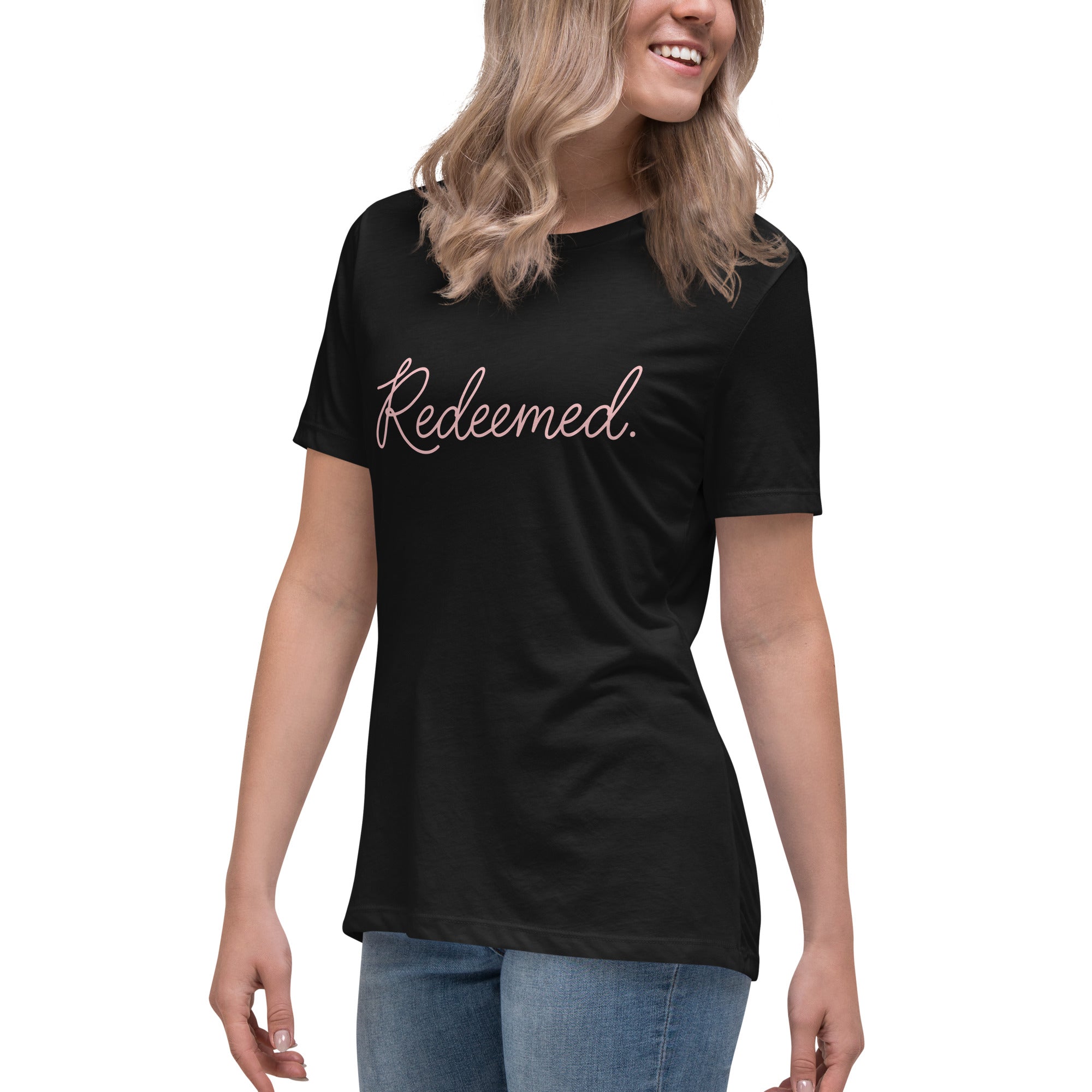 Three-quarter front left view of a blonde woman wearing a black relaxed-fit Christian t-shirt displaying the full typography design 'Redeemed.' in an elegant pink script. Lutheran women's fashion.