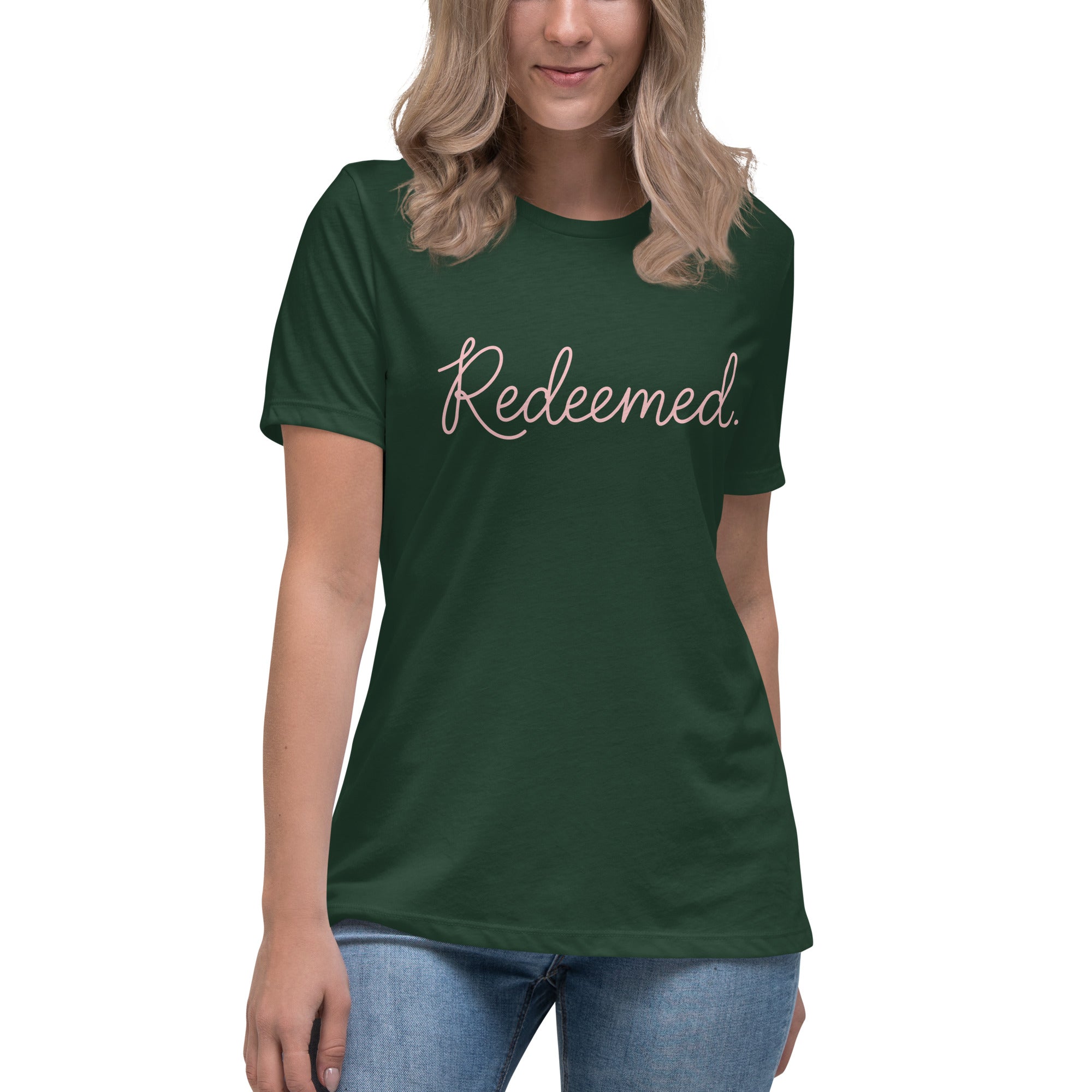A blonde woman wearing a forest green relaxed-fit Christian t-shirt featuring the word 'Redeemed.' in an elegant pink cursive font. Premium faith-based clothing for women from Phoenix Anthem.